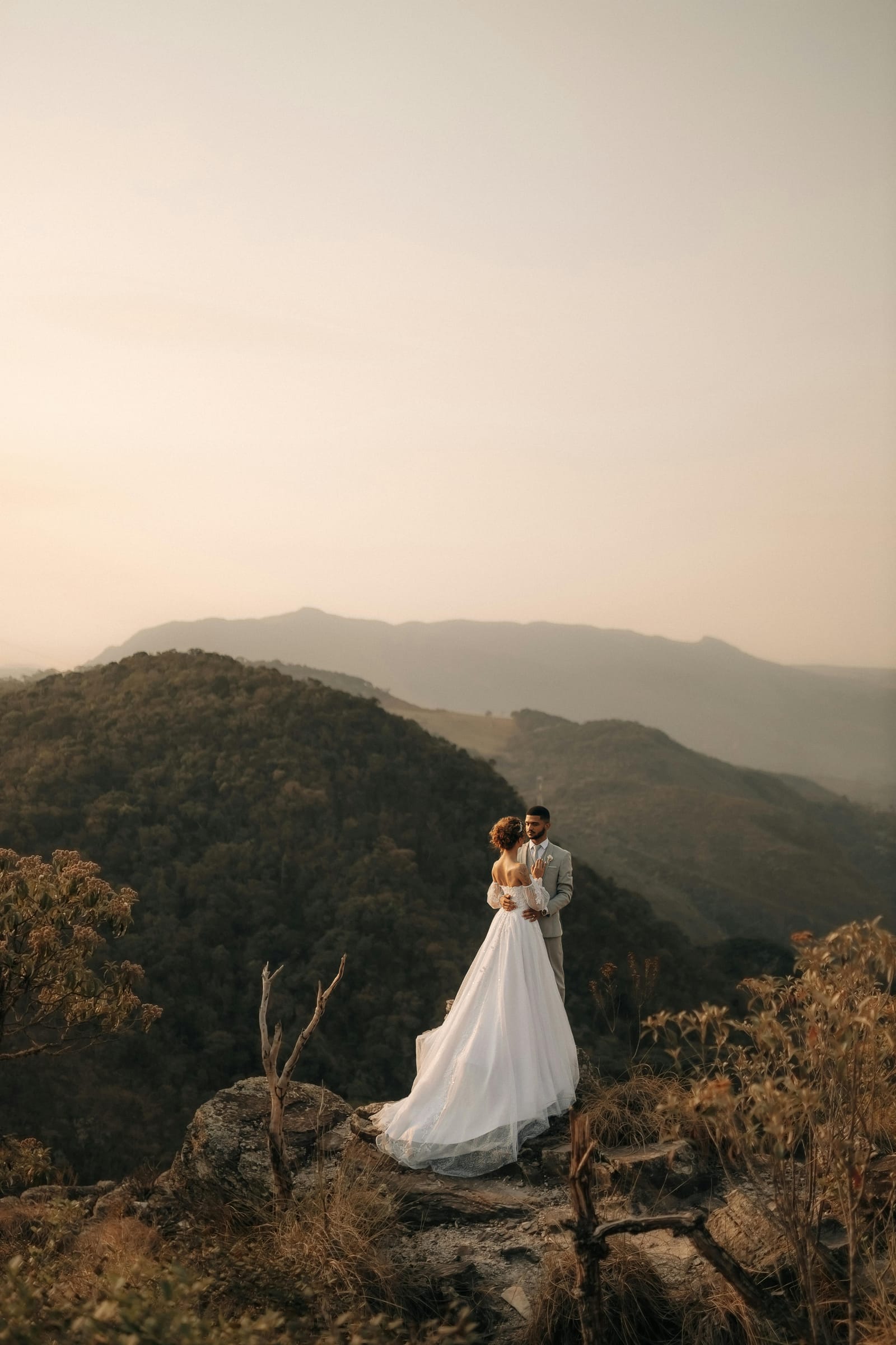 Novia de vestido blanco y novio de traje claro abrazados sobre una roca al atardecer, con montanas al fondo