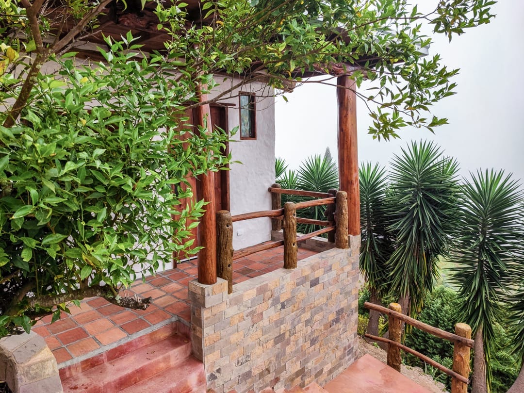 Terraza de baldosas de barro de la Cabaña Lilaila, con baranda de troncos bajo un mandarino, en Galipán