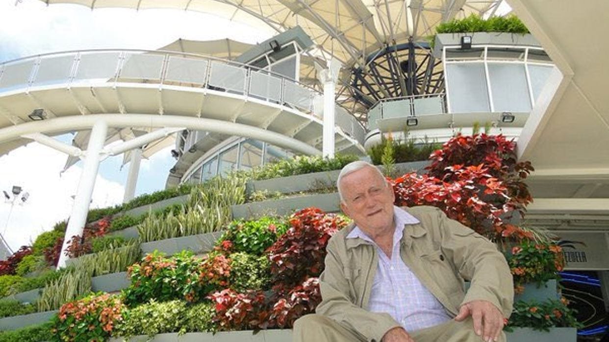 El arquitecto Fruto Vivas sentado entre jardineras floridas bajo una estructura blanca de pétalos; visitó Tarahumara